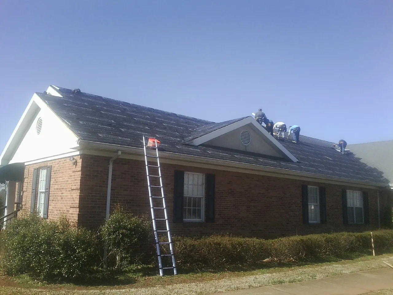 Crew tearing shingles off a brick ranch roof with a ladder set — Metal Roof Repair work by our crew in Wallis