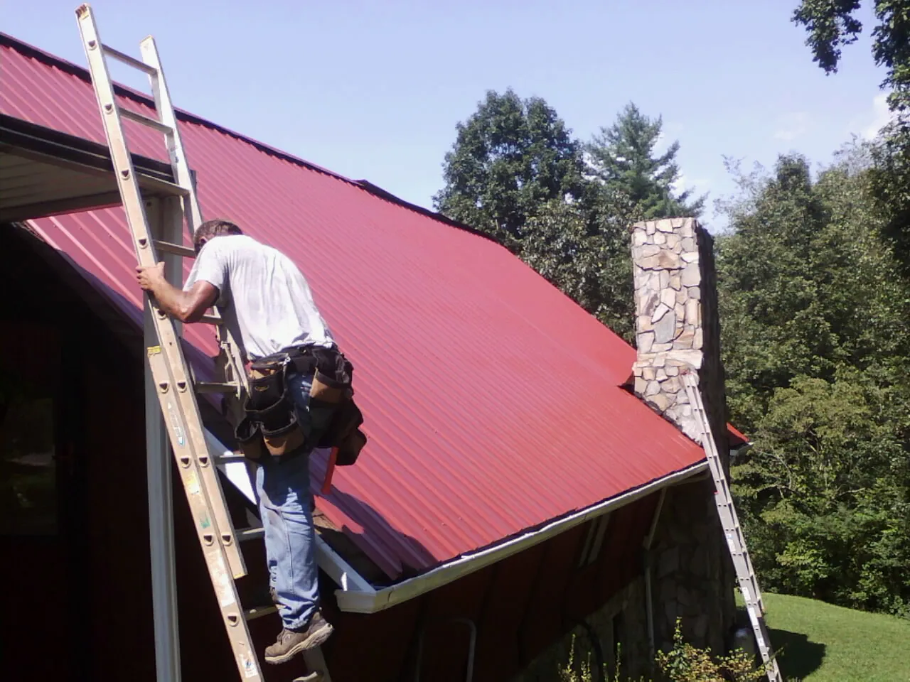 Roofer on a ladder beside a red metal roof and stone chimney — Soffit & Fascia Repair by Roofing Brothers in Wallis